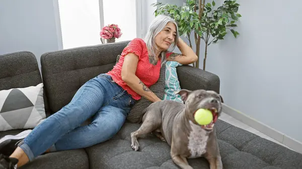Confident middle age woman, smiling happily at her pet dog, enjoying a relaxing movie time on the comfy sofa indoors