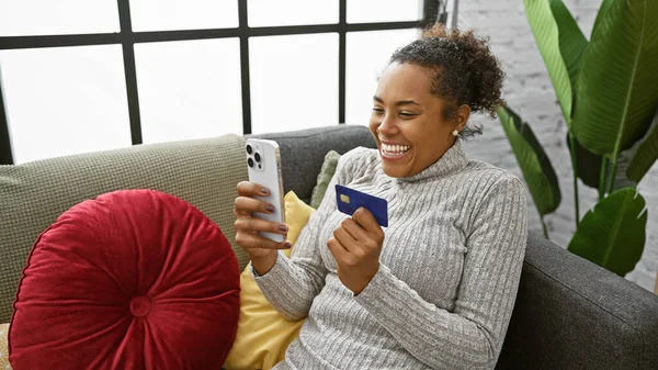Elinde kredi kartı ve akıllı telefon olan gülümseyen kadın arka planda bitkilerle koltukta oturuyor..