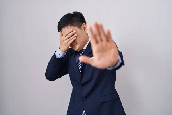 Young asian man wearing business suit and tie covering eyes with hands and doing stop gesture with sad and fear expression. embarrassed and negative concept.