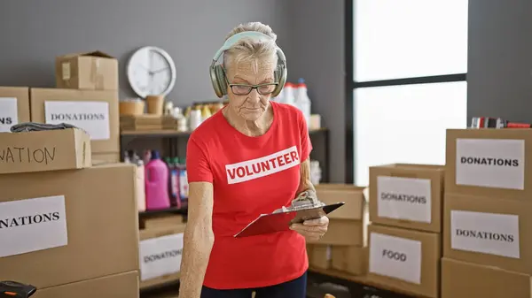 Senior grey-haired woman volunteer standing amidst community's charity center, engrossed in listening to music while checking products on checklist and reading donation report
