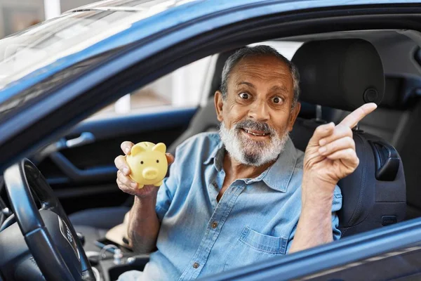 Confident elder gent with silver locks, beaming joyfully expressing optimism. outdoors in town, this one senior man points with finger at car, clutching a piggy bank. great investment or saving?