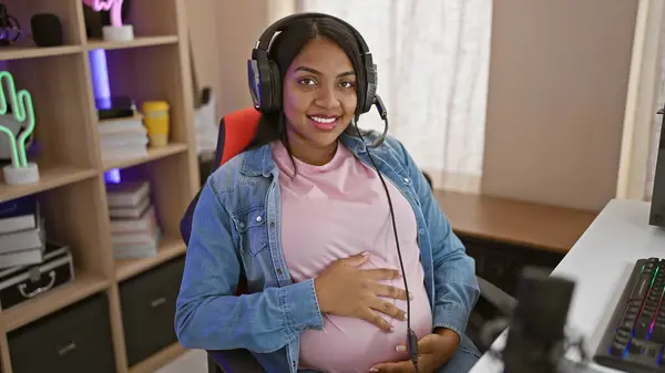 Lit from within, a young, pregnant, latina streamer beaming & touching belly while gaming in her surprisingly cozy gaming room, wearing headphones & streaming digital fun