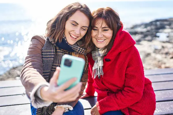 Two women mother and daughter make selfie by smartphone sitting on bench at seaside