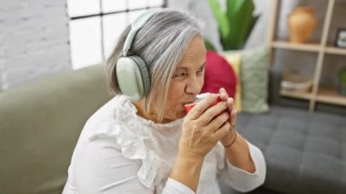 Olgun bir kadın kulaklıkla müzik dinlemekten hoşlanır ve sıcak bir oturma odasında kırmızı bir bardak tutar..