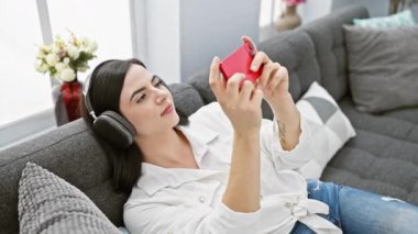 Evdeki kanepede akıllı telefon kullanan kulaklıklı, gülümseyen genç bir kadın.