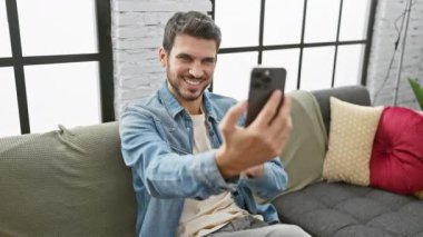 Gülümseyen genç bir adam modern bir oturma odasında selfie çekiyor, gündelik ve bağlı bir yaşam tarzını yansıtıyor..