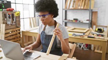 Odaklanmış bir kadın, marangozluk atölyesinde dizüstü bilgisayarla birden fazla iş yaparken odunu kontrol eder..