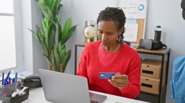 Odaklanmış Afrikalı kadın, modern ofis ortamında dizüstü bilgisayar kullanırken kredi kartı tutuyor, online alışveriş veya ödeme kavramını yansıtıyor.