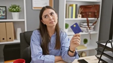 Düşünceli genç bir İspanyol kadın modern ofis ortamında kredi kartı tutuyor, profesyonellik ve düşünce dolu..