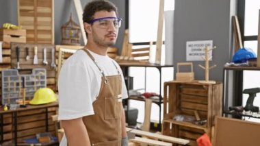 Güvenlik gözlüğü takan sakallı ve önlüklü genç adam iyi organize edilmiş bir marangozluk atölyesinde kollarını kavuşturarak duruyor..