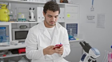 Laboratuvar ortamında akıllı telefon kullanan laboratuvar önlüklü gülümseyen genç bir adam. Arka planda ekipman ve beyaz dolaplar var..