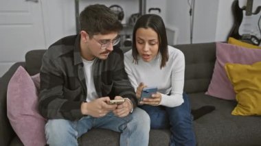 Bir erkek ve bir kadın, evlerinde modern bir çifti örnek alarak, rahat bir oturma odasında akıllı telefonlar kullanarak bir kanepeye yakın otururlar..