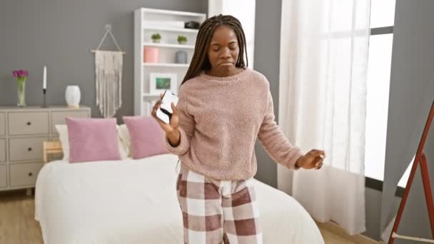 一个快乐的非洲女人 头戴辫子 手握智能手机 在舒适的卧室里跳舞 展示着悠闲的家庭生活方式 — 图库视频影像