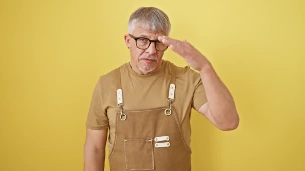 眼鏡に真剣な中年の灰色の髪の男性は 手で目を覆い 悲しみと拒絶の目立たない表情を体現し 厳しい黄色の背景に対して一人で立っています — ストック動画