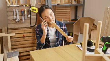 Marangozluk atölyesinde akıllı telefonla konuşan Asyalı genç bir kadın ahşabı teftiş ediyor..