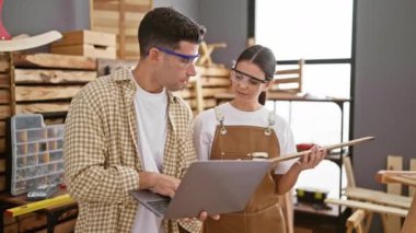 Güvenlik gözlüğü takan bir erkek ve kadın, ahşap atölyesinde iş birliği yapıyor, proje detaylarını dizüstü bilgisayarla tartışıyorlar..