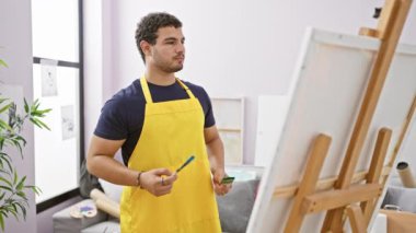 Parlak bir stüdyoda tuvale odaklanmış genç bir adam sarı bir önlük giyiyor ve elinde sanat malzemeleri tutuyor..