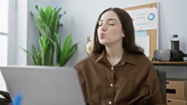 Kahverengi bluzlu genç beyaz kadın, modern ofis ortamında dizüstü bilgisayarının başında neşeli bir yüz ifadesi sergiliyor..