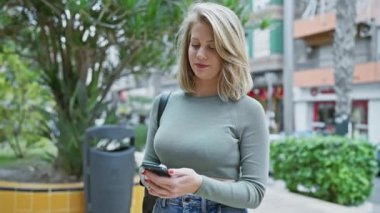 Genç sarışın bir kadın, işlek bir şehir caddesinde akıllı telefonunu kullanarak şehir merkezindeki modern bağlantıları vurguluyor..