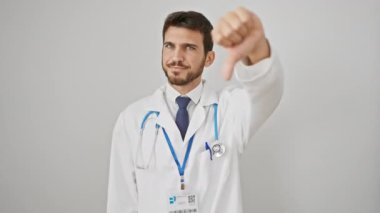 Beyaz önlüklü kızgın, genç, ispanyol bir adam, baş parmaklarını aşağı gösteriyor. Kötü titreşimler ve steteskoplu mutsuz bir doktorun ifadesi beyaz duvarda izole edilmiş. hiç hoşlanmadım.!