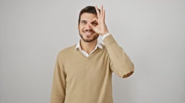 Yakışıklı, genç İspanyol bir adam, gözlerinde bir mutlulukla, parmaklarıyla başarılı bir 'ok' işareti yapıyor, beyaz bir arka planda neşeli bir şekilde duruyor, modaya uygun bir süveter giyiyor..
