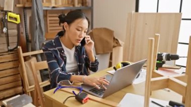 Genç Asyalı kadın dizüstü bilgisayarda çalışıyor ve marangozluk atölyesinde telefonla konuşuyor..
