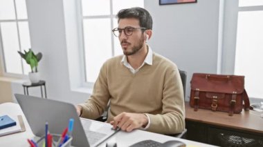 Ofiste dizüstü bilgisayar ve evrak çantasıyla rahatlayan gözlüklü profesyonel İspanyol adam.