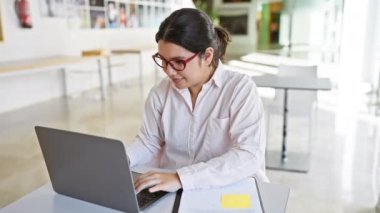 Odaklanmış İspanyol bir kadın modern bir ofiste çalışıyor, dizüstü bilgisayarında yazıyor ve notlar yazıyor..