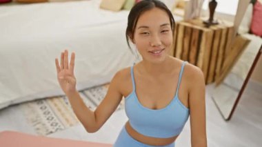 Neşeli genç Çinli kadın parmaklarıyla 'dört' sayıyor, yatak odasındaki yoga minderinden yukarıya doğru neşeyle işaret ediyor, pozitif yüz ifadesi gösteriyor.