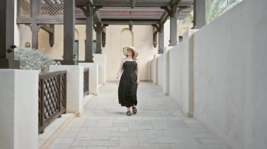 Siyah elbiseli ve hasır şapkalı gülümseyen bir kadın Dubai 'deki Madinat Jumeirah' ın geleneksel mimarisinde yürüyor..