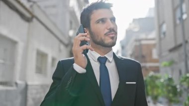 Genç bir işadamı güneşli bir şehir caddesinde akıllı telefon kullanıyor. İletişim ve modern yaşam tarzını çağrıştırıyor..