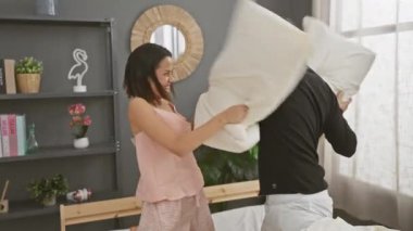 Mutlu çift rahat, modern bir yatak odasında neşeli bir yastık kavgası yapıyor, neşe, sevgi ve evde rahat bir yaşam tarzı sergiliyor..