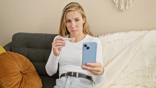 金髪の女性は 屋内の灰色のソファに座ってスマートフォンを保持しながら デジタル温度計で彼女の温度をチェックします — ストック動画