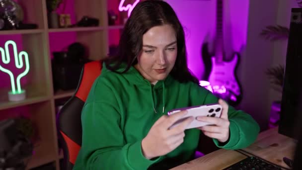 若い女性がネオンライトの部屋で彼女の携帯電話でビデオゲームをプレイし 喜びと集中を表現する — ストック動画