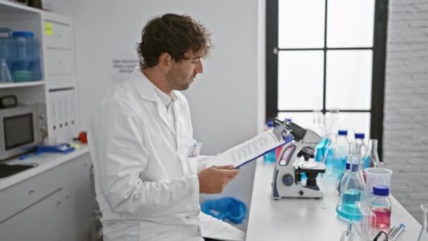 穿着实验室外套的西班牙裔男子在明亮的实验室用显微镜和化学仪器检查剪贴板 — 图库视频影像