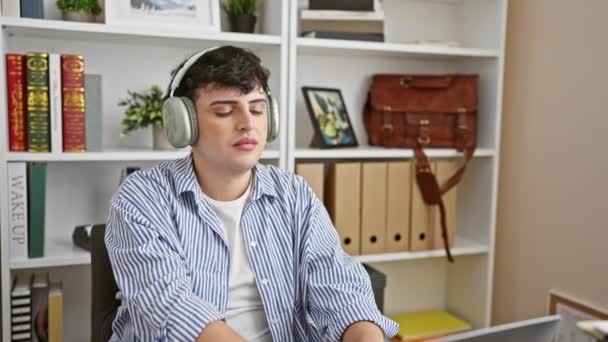 在一个充满书架和书籍的现代化办公室里 一个带着耳机的轻松的年轻人享受着音乐的休息 — 图库视频影像