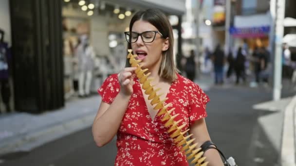 美しいヒスパニックの女性が喜んでクランチを食べる タケシタ通りのスティック上の美味しいチップ 日本の都市でジャンクフード愛好家 スポーツメガネと陽気な笑顔を旅行 — ストック動画