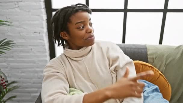 快乐的年轻黑人妇女 头发蓬乱 坐在舒适的沙发上 微笑着 用她的手指和手 带着一种自然的喜悦的表情 玩世不恭地抬起头 — 图库视频影像