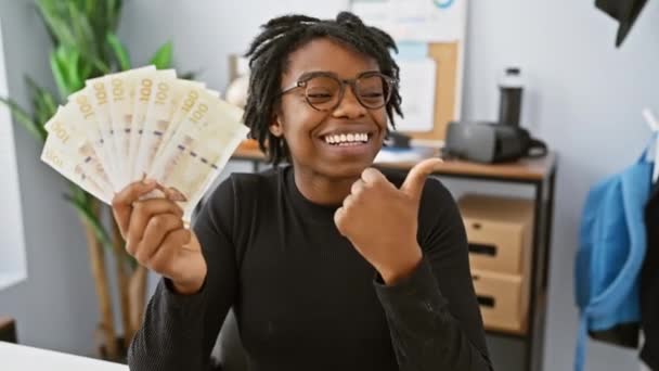 オフィスのドレッドロックを持つ陽気な若い黒人女性は 喜んで側を指し デンマークのクローン紙幣を保持し 開いた口と親指で大きな自信を持って笑顔を点滅し ジェスチャーを上げます — ストック動画