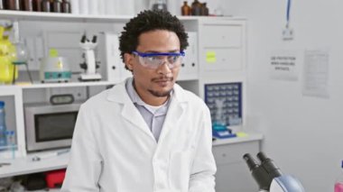 Neşeli, Afro-Amerikan laboratuvar bilim adamı, güvenlik gözlüğü takan, mutlu bir şekilde bir parmağını havaya kaldıran, modern araştırma merkezinde kendine güvenen bir gülümsemeyle aklına gelen zeki bir fikir..