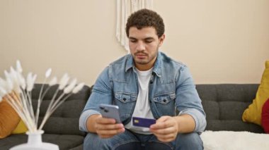 Modern bir oturma odasında elinde akıllı telefon ve kredi kartı olan kafası karışmış bir adam..