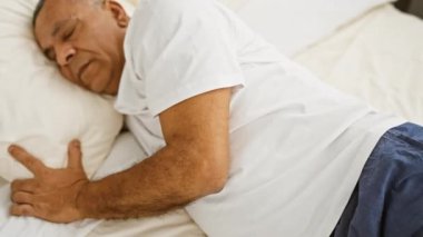 Beyaz tişörtlü ve donanma pantolonlu orta yaşlı bir İspanyol iyi ışıklandırılmış bir yatak odasında uyur ve yastığı kucaklar..