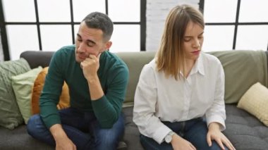 Bir erkek ve kadın uzaktan koltuğa oturur, modern bir oturma odasında gerginlik ve kopukluk ifade ederler..