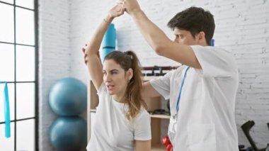 Klinikteki erkek bir fizyoterapist, içerideki rehabilitasyon egzersizinde gülümseyen bir kadına rehberlik eder..