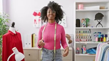 Kolları çaprazlanmış, modern atölyesinde dikilmiş, etrafı dikiş malzemeleri ve mankenlerle çevrili kendinden emin Afrikalı bir kadın..