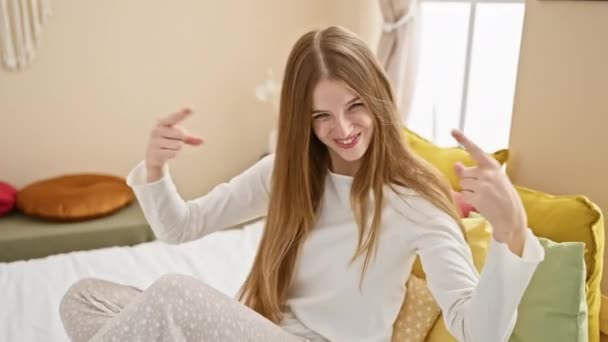 自信と誇りを持ってパジャマの若いブロンドの女性は 喜んでベッドルームのベッドに座って 陽気に見える自己を指しています — ストック動画