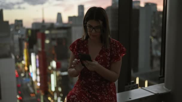 写真を魅了し デジタル会話で失われたメガネの美しいヒスパニック女性 東京のパノラマの中に座っている間 スマートフォンにタイピング — ストック動画