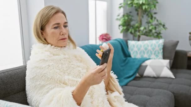 成熟したブロンドの女性はリビングルームのソファー 携帯電話にハドリードしながら懸念を持って薬瓶を検査します — ストック動画