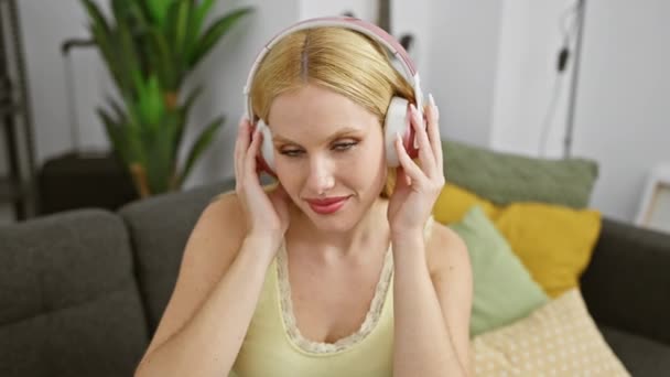金发女人在舒适的客厅里用耳机听音乐 在家里展示休闲和放松 — 图库视频影像