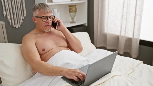 成熟した男 屋内ベッドで座っている間 ラップトップと電話の会話とマルチタスク — ストック動画